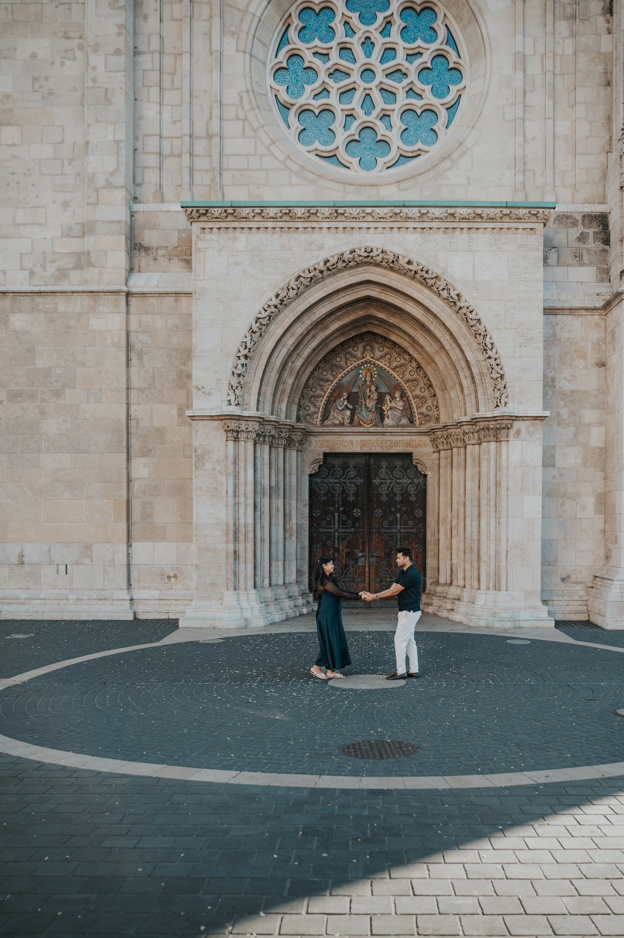 The couple standing below the large carved portal and blue rose window at Matthias Church during a couple photoshoot in Budapest.