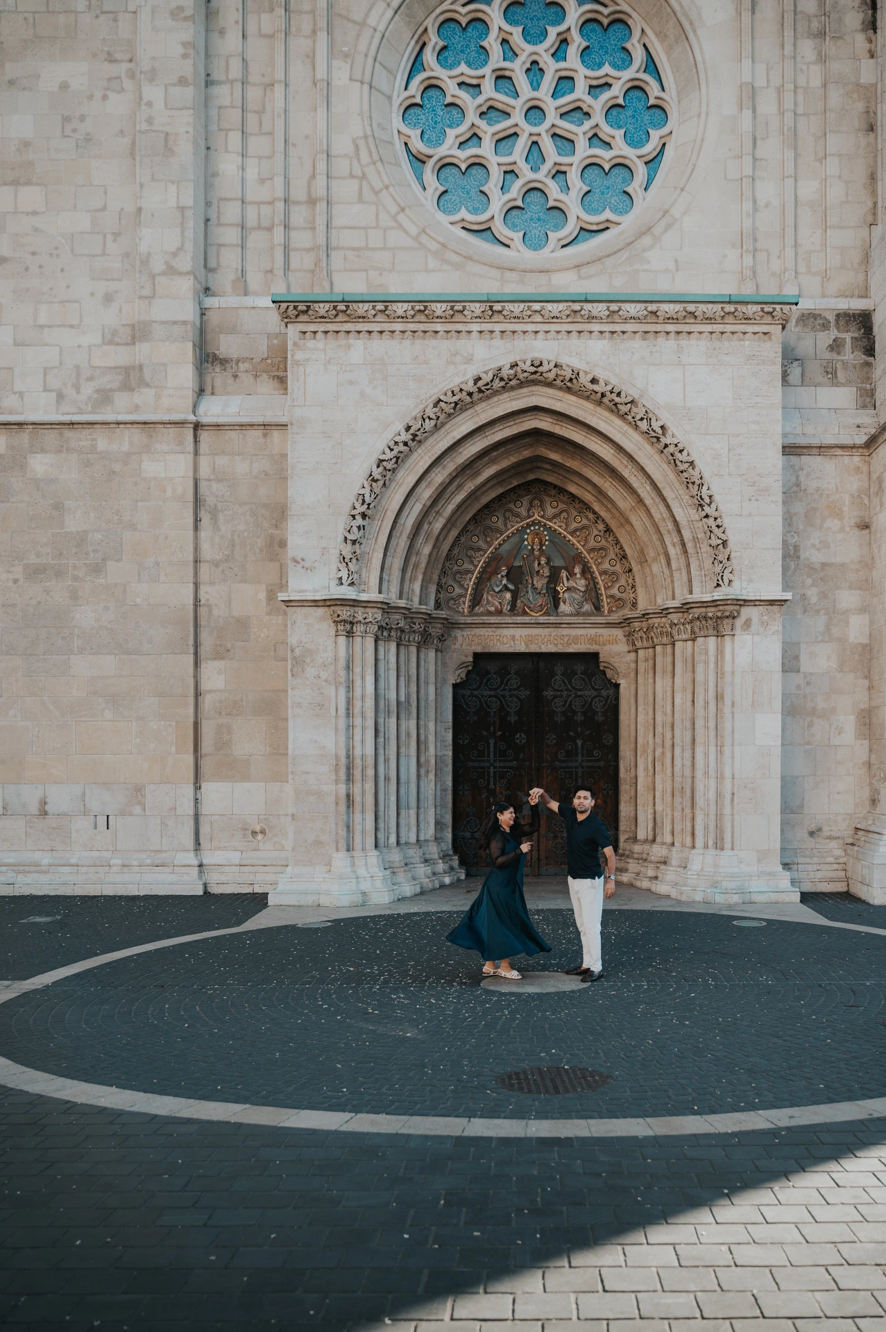 The couple turning together in the circular paved area beneath Matthias Church’s blue rose window during a couple photoshoot in Budapest.