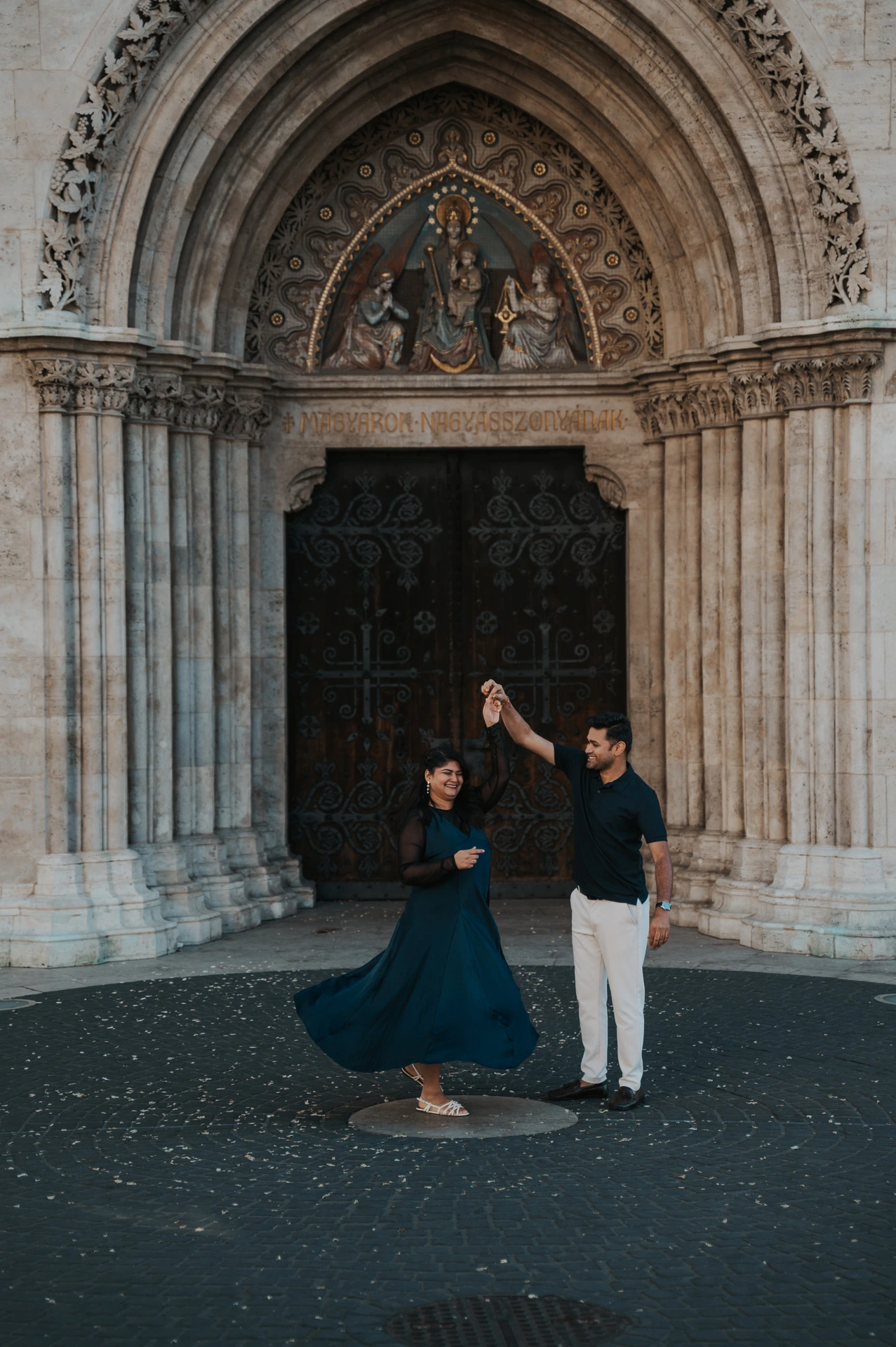 The woman’s teal dress swirling as her partner raises her hand before Matthias Church’s carved portal during a couple photoshoot in Budapest.