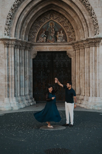 Budapest couple photoshoot: a swirling teal dress beside the church portal The woman’s teal dress swirling as her partner raises her hand before Matthias Church’s carved portal during a couple photoshoot in Budapest.