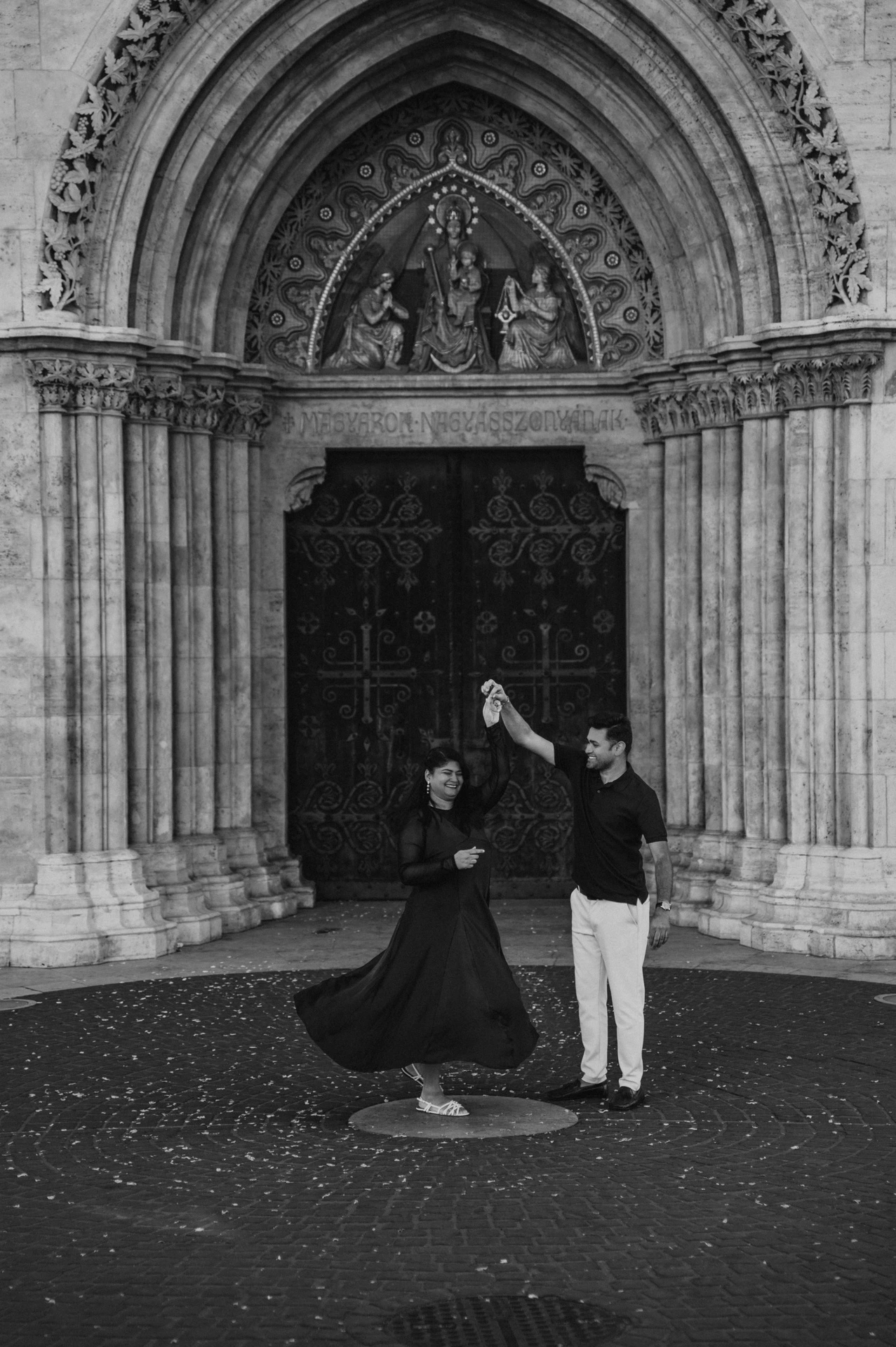 Black-and-white photograph of the couple turning together with the woman’s dress sweeping across the paving before Matthias Church during a couple photoshoot in Budapest.