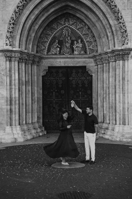 Budapest couple photoshoot: a dress in motion before the carved church portal in black and white Black-and-white photograph of the couple turning together with the woman’s dress sweeping across the paving before Matthias Church during a couple photoshoot in Budapest.