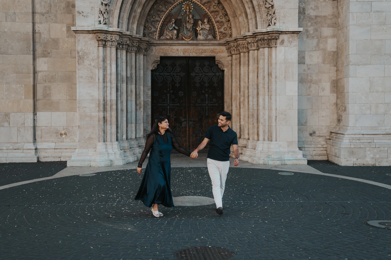 Budapest couple photoshoot: walking hand in hand from the church portal A landscape portrait of the couple walking hand in hand away from Matthias Church’s carved portal during a couple photoshoot in Budapest.