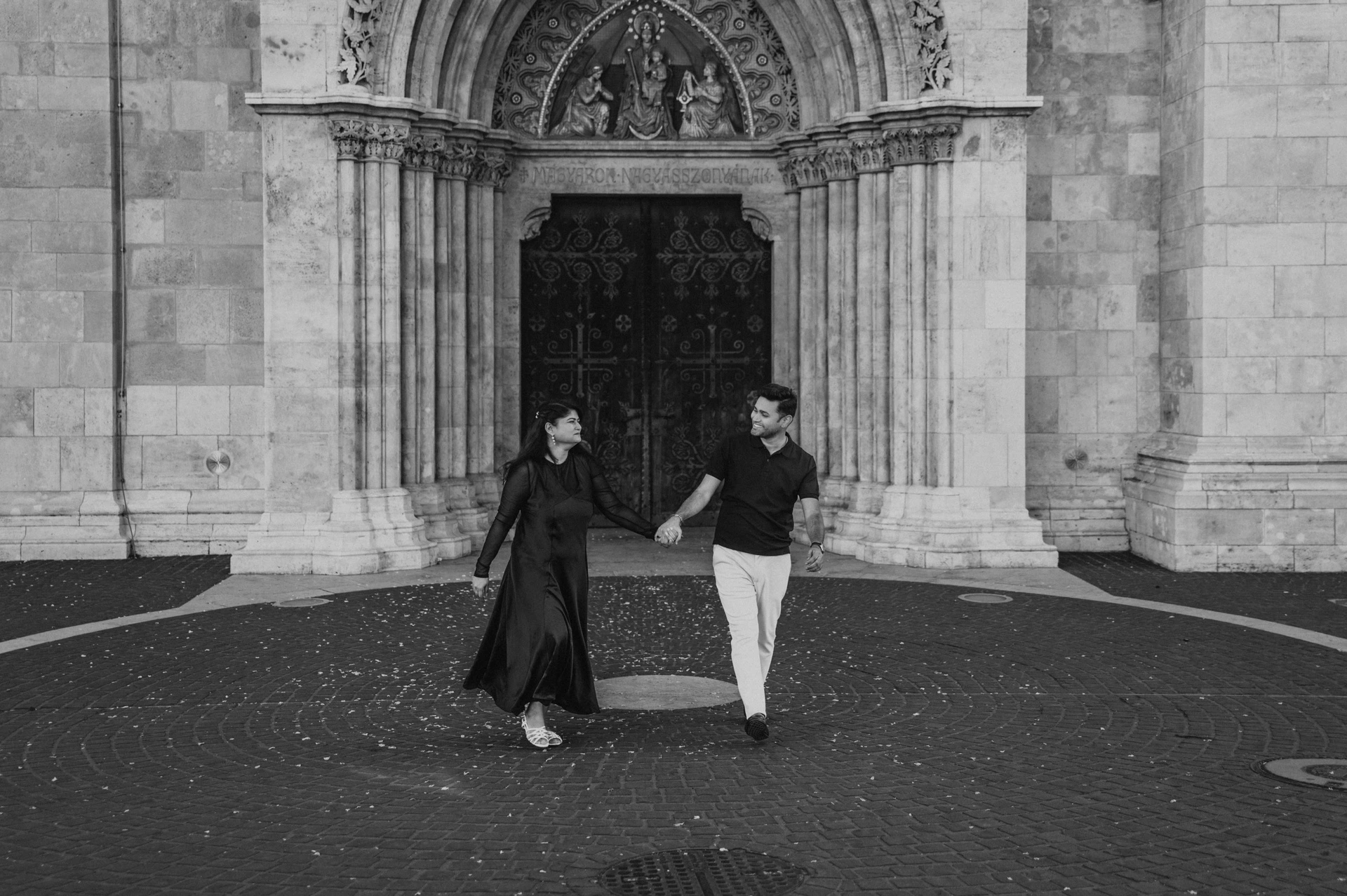 Black-and-white photograph of the couple walking together across the paving in front of Matthias Church’s large carved doorway during a couple photoshoot in Budapest.