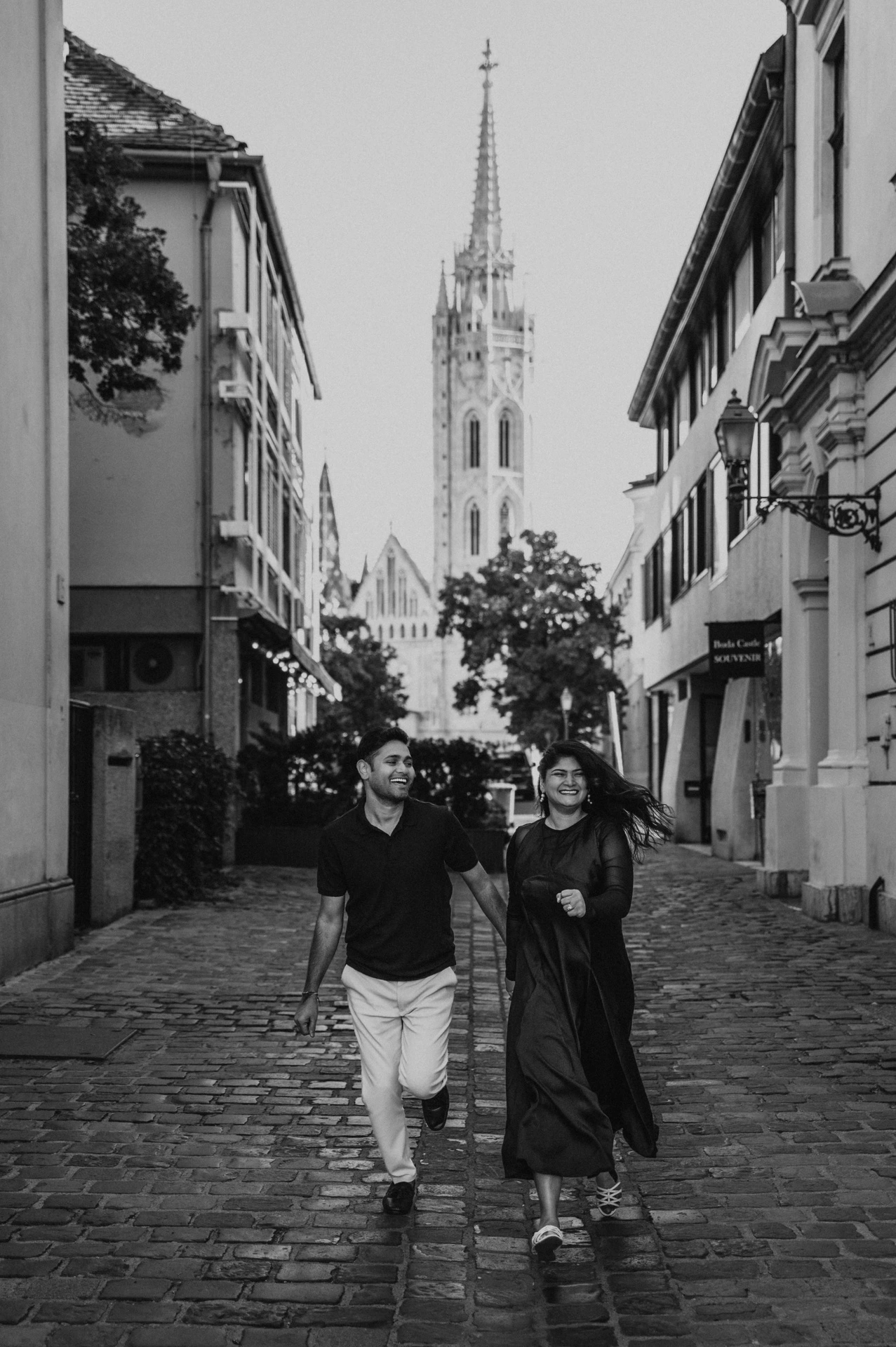 Black-and-white photograph of the couple walking hand in hand along a cobbled street framed by buildings and the church spire during a couple photoshoot in Budapest.