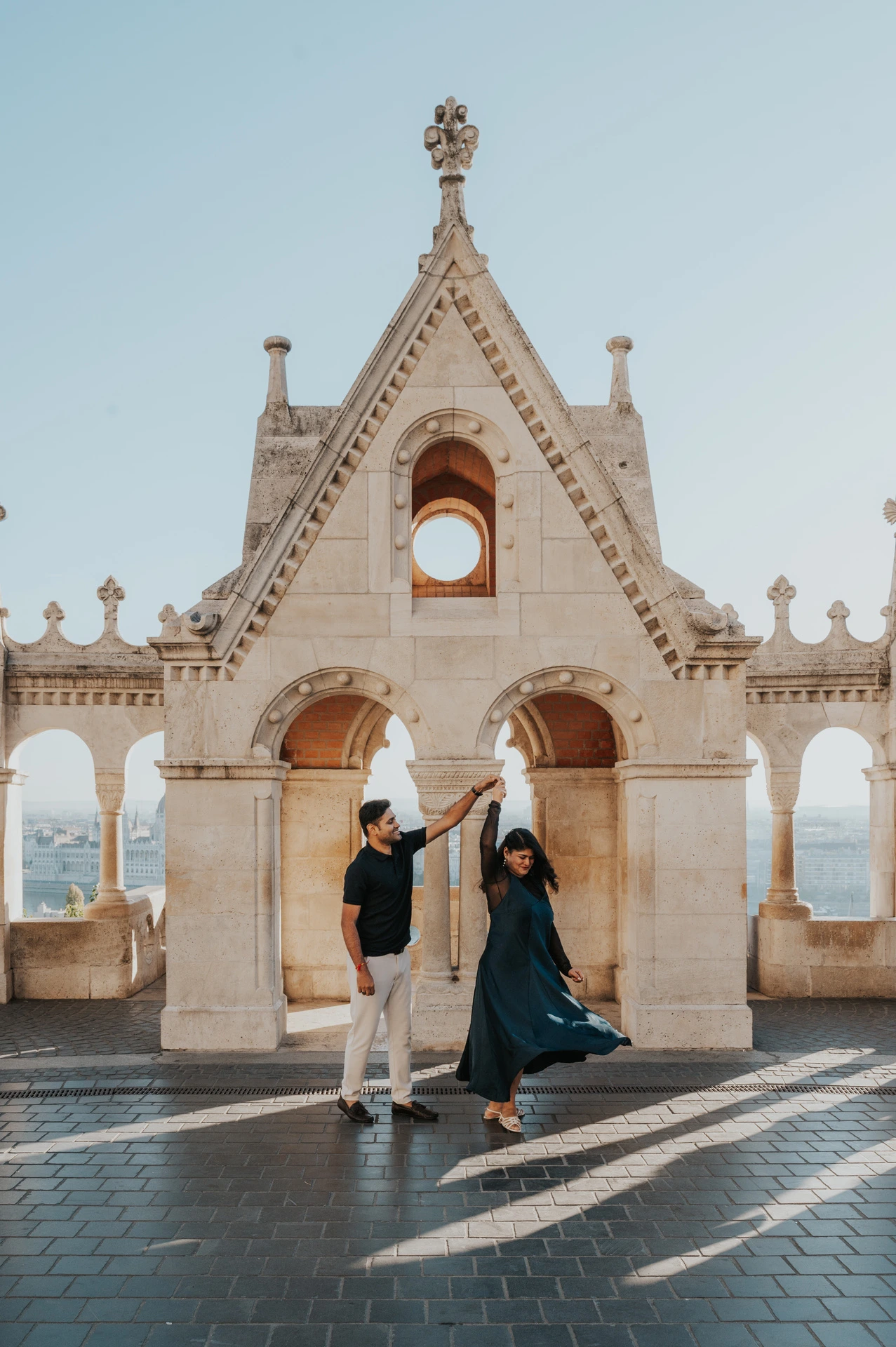 The woman’s teal dress sweeping outward as the couple turn beneath the decorative Bastion gable during a couple photoshoot in Budapest.