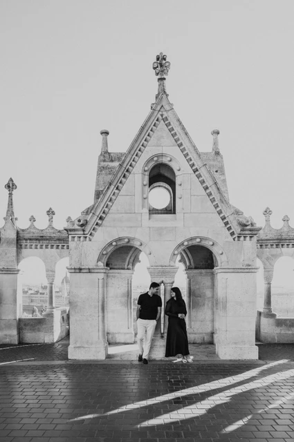 Budapest couple photoshoot: the gabled entrance and patterned shadows in black and white Black-and-white photograph of the couple standing under the ornate Bastion gable with long shadows across the paving during a couple photoshoot in Budapest.