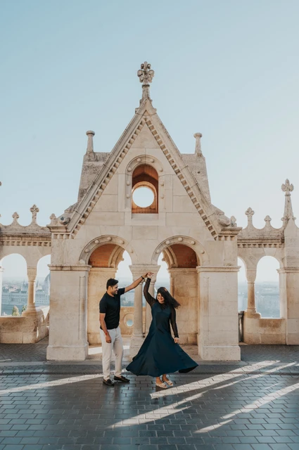 Budapest couple photoshoot: a teal dress turning in the Bastion forecourt The man raising his partner’s hand as her teal dress turns in front of an ornate Bastion gable during a couple photoshoot in Budapest.