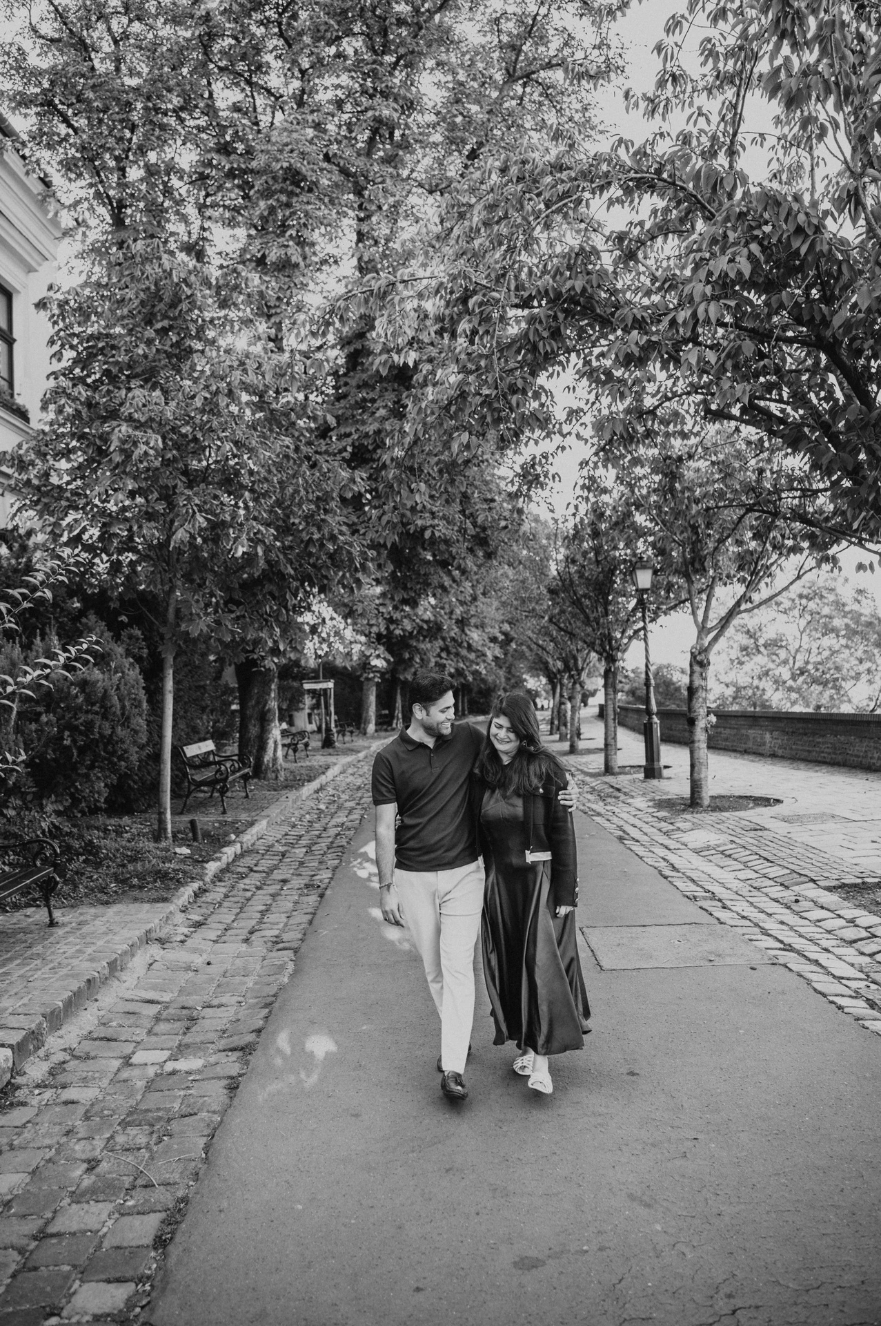 Black-and-white photograph of the couple walking together beneath the trees on Tóth Árpád promenade during a couple photoshoot in Budapest.