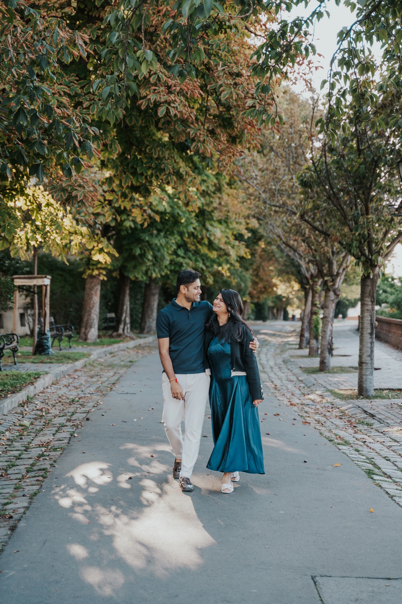 The couple walking together beneath green and golden leaves on Tóth Árpád promenade during a couple photoshoot in Budapest.