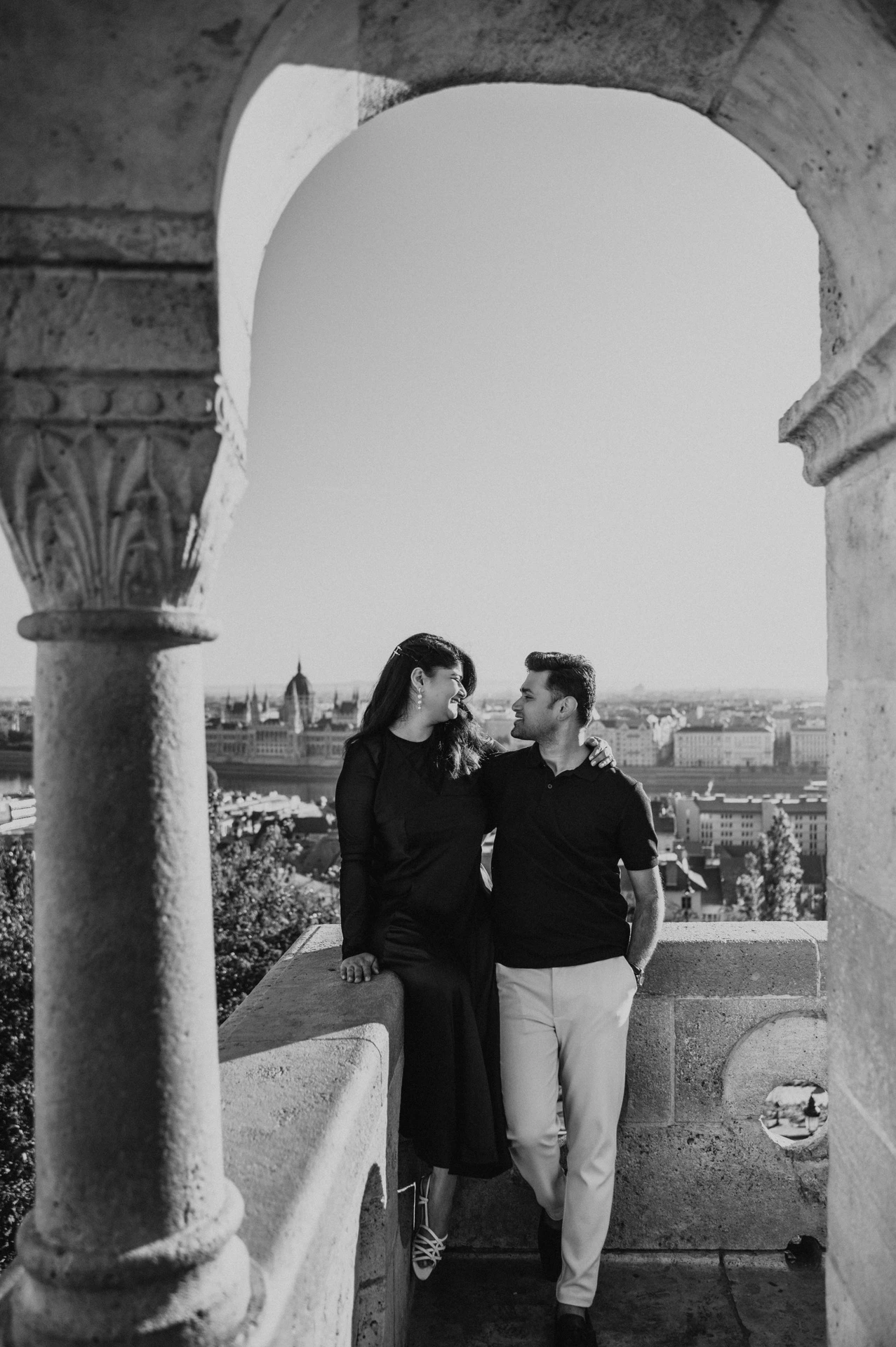 Black-and-white photograph of the couple leaning towards each other beneath a terrace arch at Fisherman’s Bastion during a couple photoshoot in Budapest.