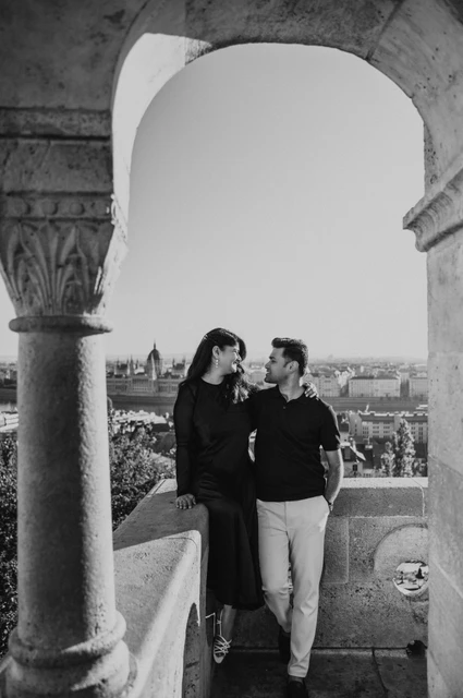 Budapest couple photoshoot: leaning close beneath a terrace arch in black and white Black-and-white photograph of the couple leaning towards each other beneath a terrace arch at Fisherman’s Bastion during a couple photoshoot in Budapest.