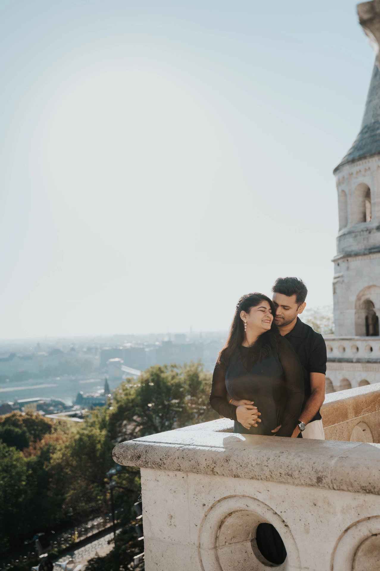 The couple sitting close beside a carved stone balustrade with a Bastion tower and city beyond during a couple photoshoot in Budapest.