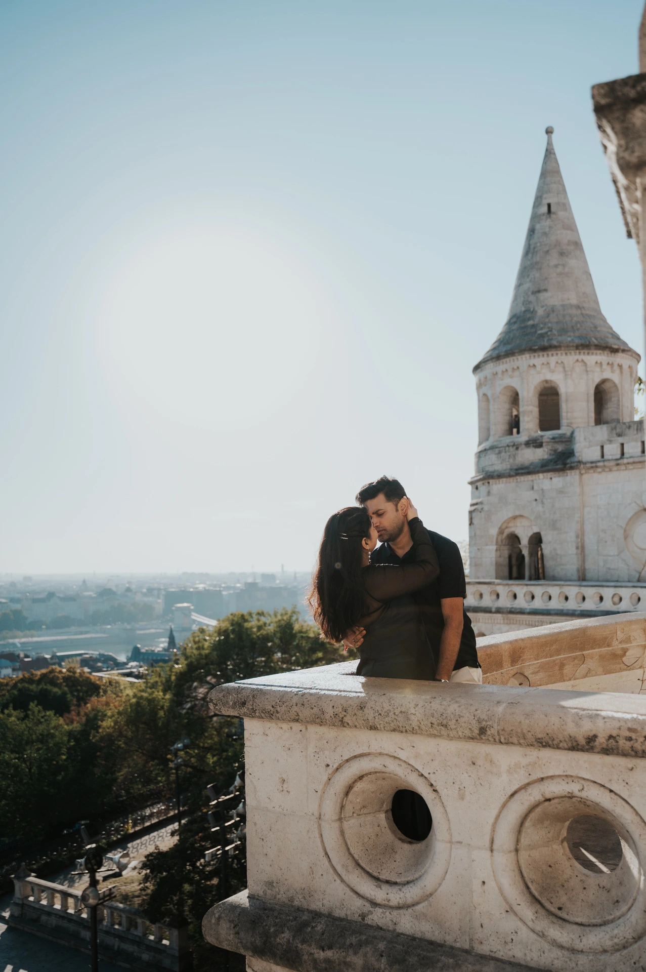 The couple seated beside the carved stone balustrade with a pointed Bastion tower behind them during a couple photoshoot in Budapest.