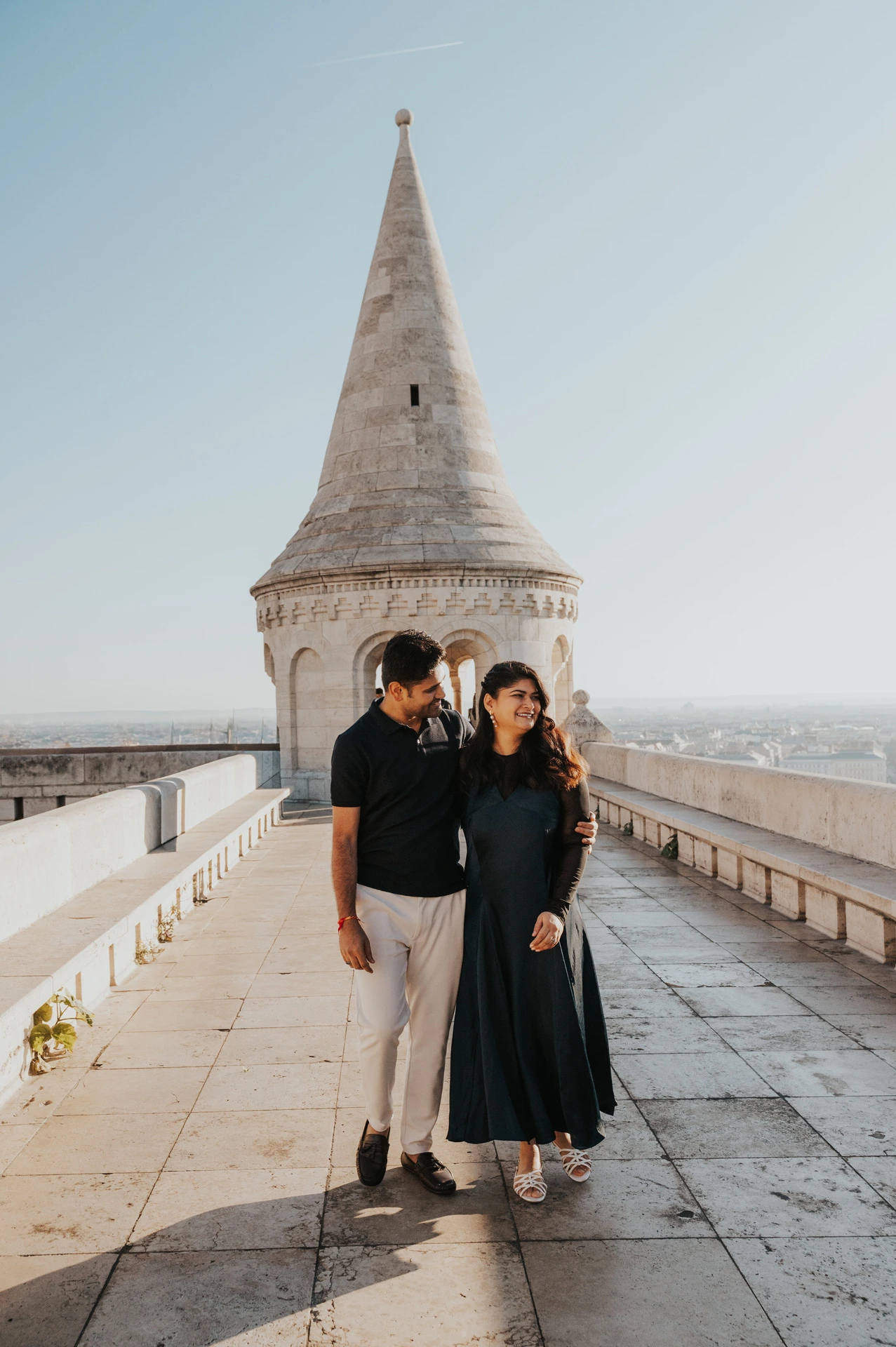 The couple walking together along the stone terrace beneath a pointed tower at Fisherman’s Bastion during a couple photoshoot in Budapest.