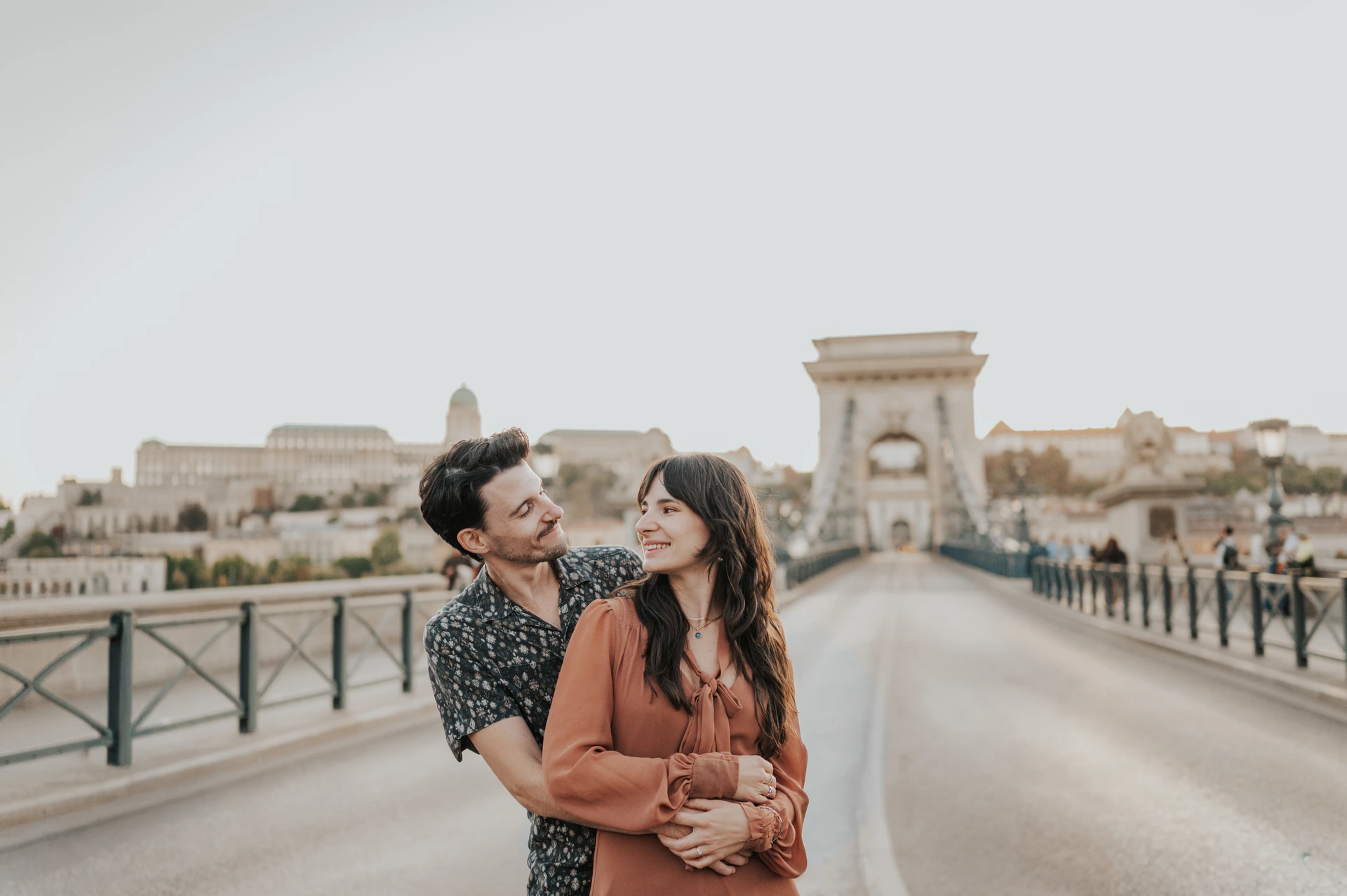 Couple embracing on the Chain Bridge in Budapest, with the bridge tower and Buda Castle in the background.