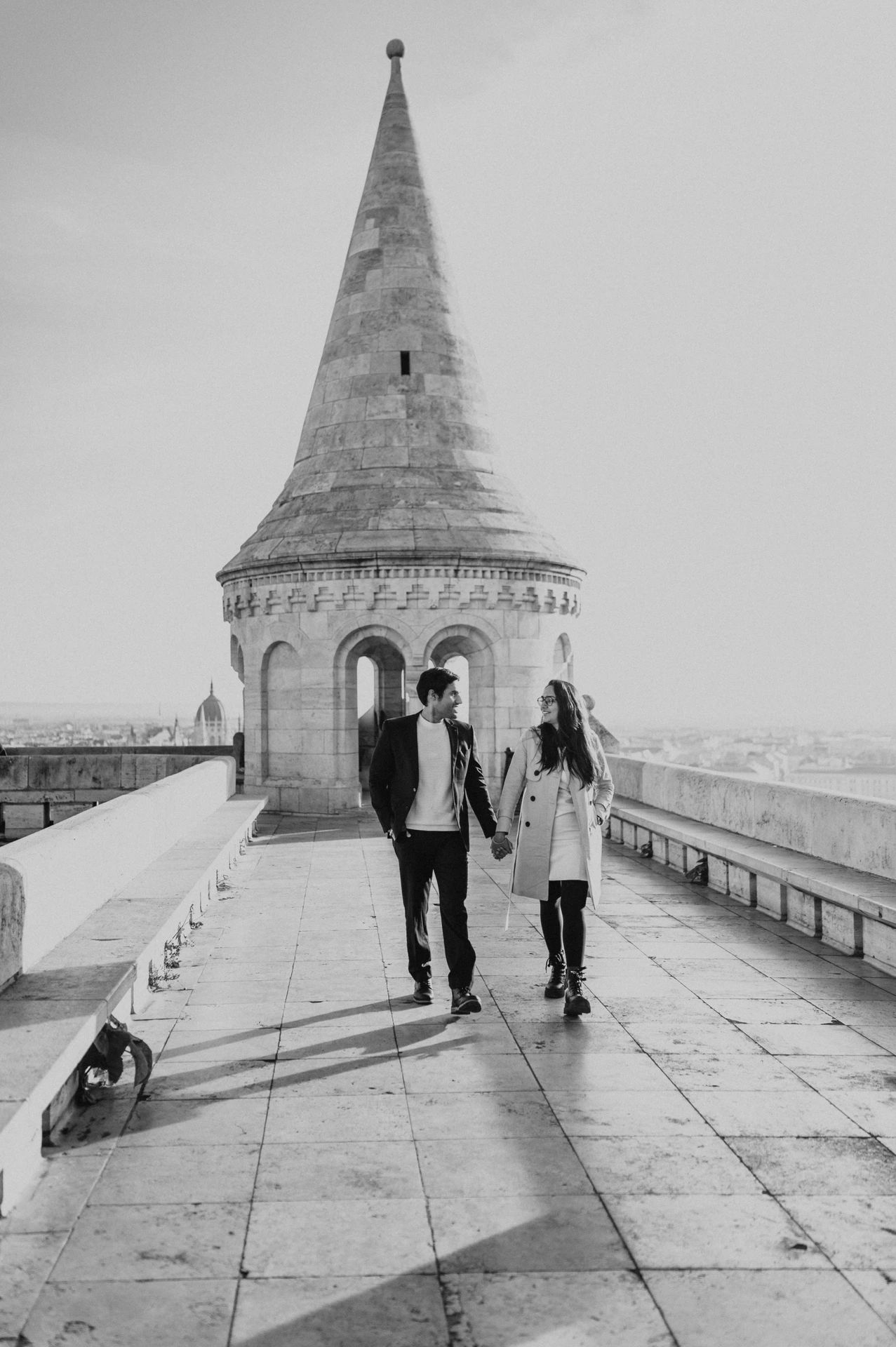 Black and white photograph of a couple walking hand in hand by a Fisherman's Bastion tower during their photoshoot in Budapest