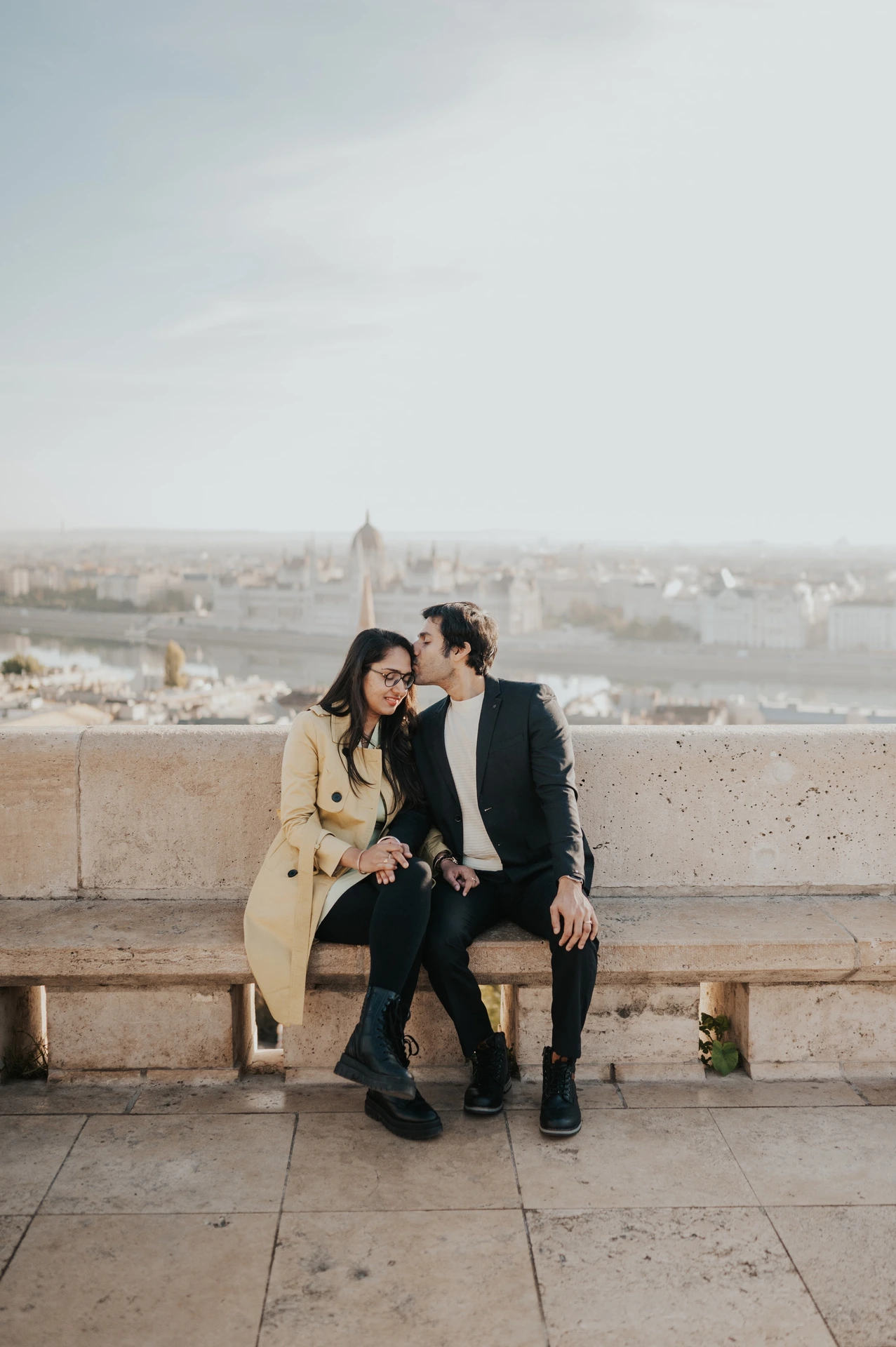 Couple sitting together by the Fisherman's Bastion balustrade during their photoshoot in Budapest, with the city behind them
