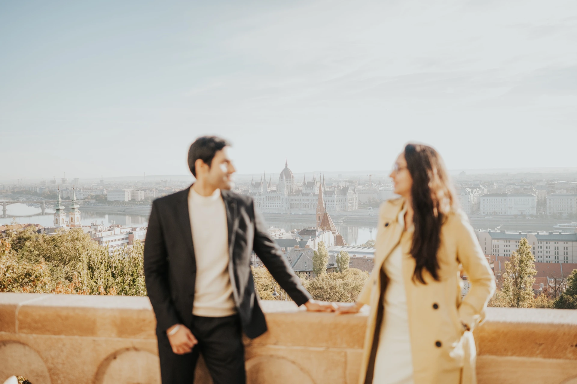 Wide photograph of a couple holding hands at Fisherman's Bastion during their Budapest photoshoot, with the Parliament building behind them