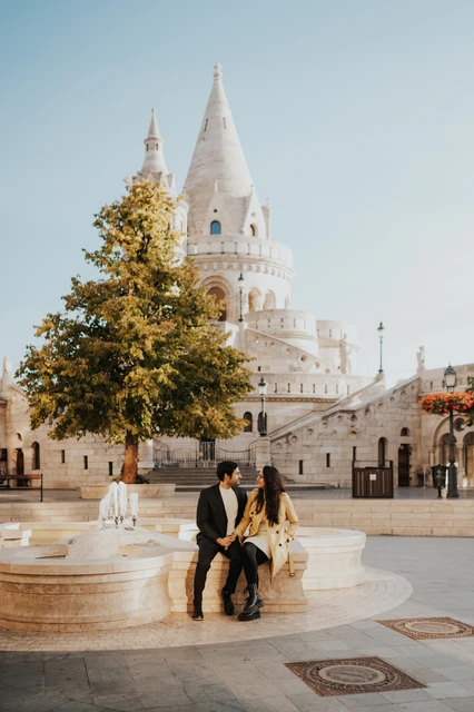 Couple photoshoot in Budapest near Fisherman's Bastion with the church spire behind them Couple sitting on a curved stone ledge in the square near Fisherman's Bastion during their photoshoot in Budapest