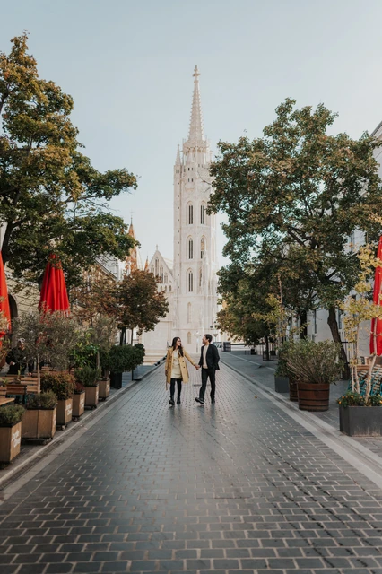 Morning couple photoshoot in Budapest on a cobbled street near Fisherman's Bastion Couple walking hand in hand along a cobbled street lined with trees near Fisherman's Bastion during their morning photoshoot in Budapest