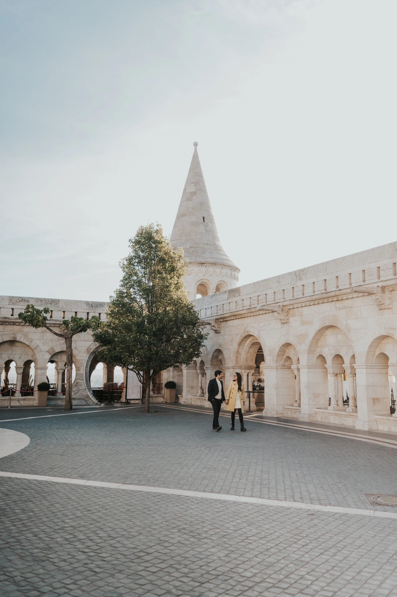 Wide portrait of a couple standing in the square with the towers and arcades of Fisherman's Bastion behind them during their Budapest photoshoot