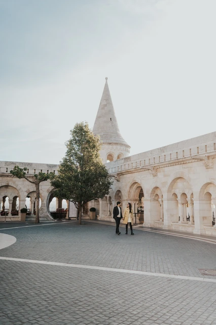 Couple photoshoot in Budapest with Fisherman's Bastion in the backdrop Wide portrait of a couple standing in the square with the towers and arcades of Fisherman's Bastion behind them during their Budapest photoshoot