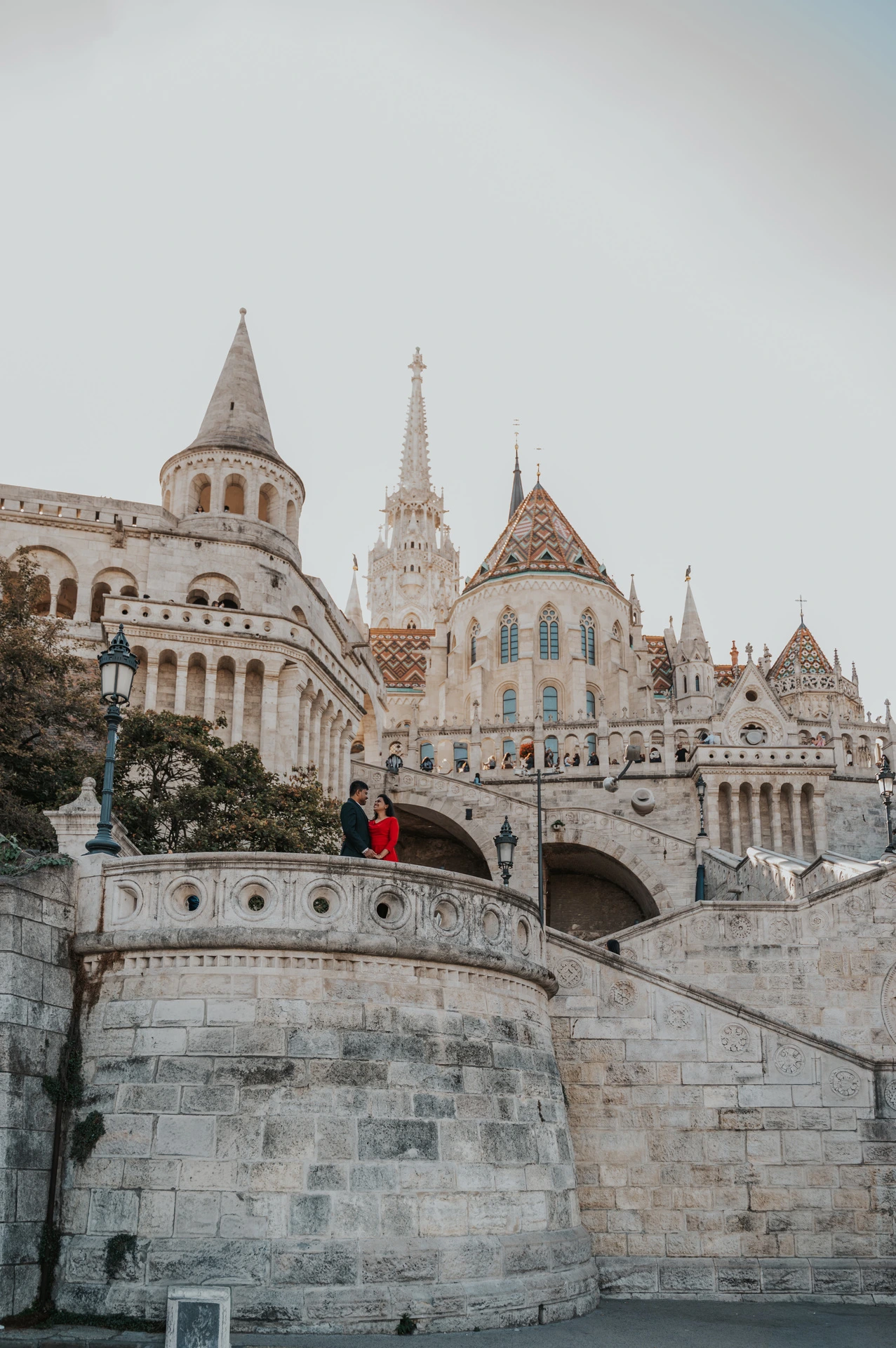 The couple appearing on a rounded terrace beneath Fisherman’s Bastion’s tower, with a patterned church roof beyond, during their Budapest photoshoot.