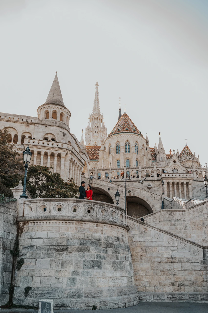 A tower and tiled roof above a Budapest couple photoshoot The couple appearing on a rounded terrace beneath Fisherman’s Bastion’s tower, with a patterned church roof beyond, during their Budapest photoshoot.
