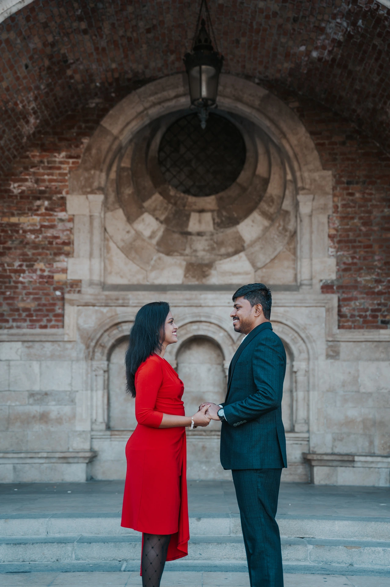 The couple holding hands and facing one another beneath a brick-and-stone arch with a round window during their couple photoshoot in Budapest.