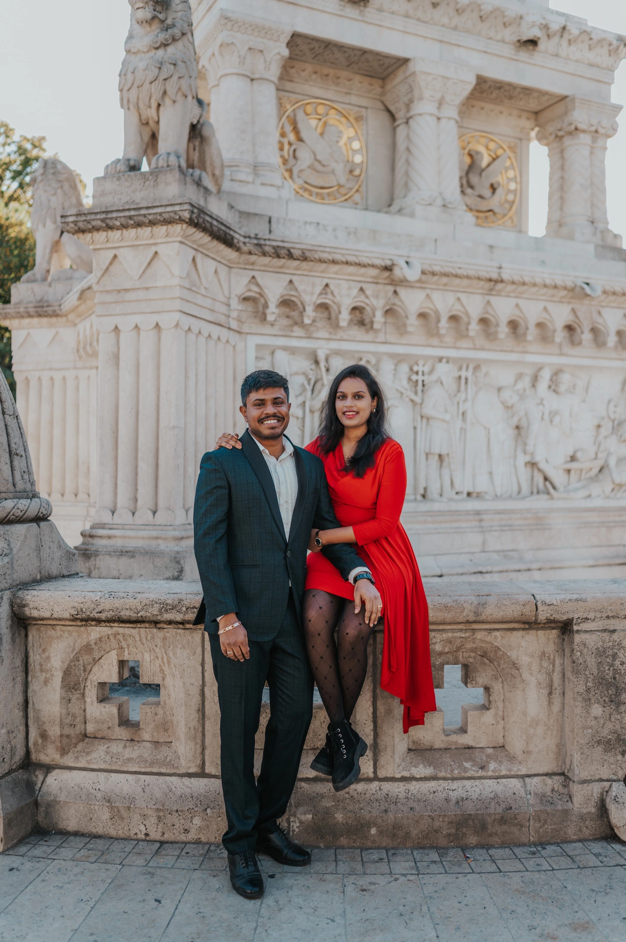 The couple facing the camera beside a carved stone monument, with the woman seated on the low wall during their Budapest photoshoot at Fisherman’s Bastion.