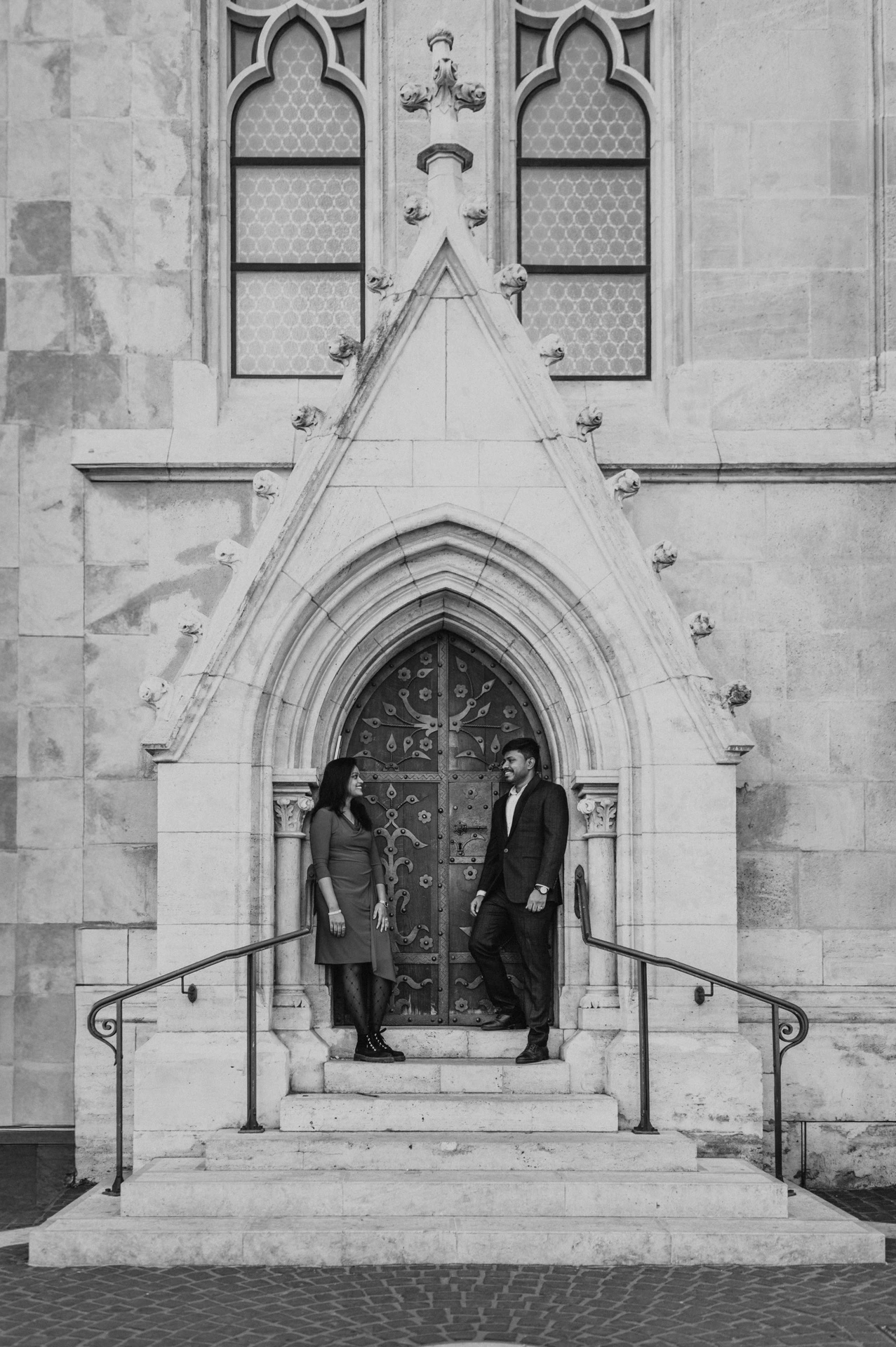 Black-and-white portrait of the couple standing on the church steps beneath a tall Gothic doorway during their Budapest photoshoot.