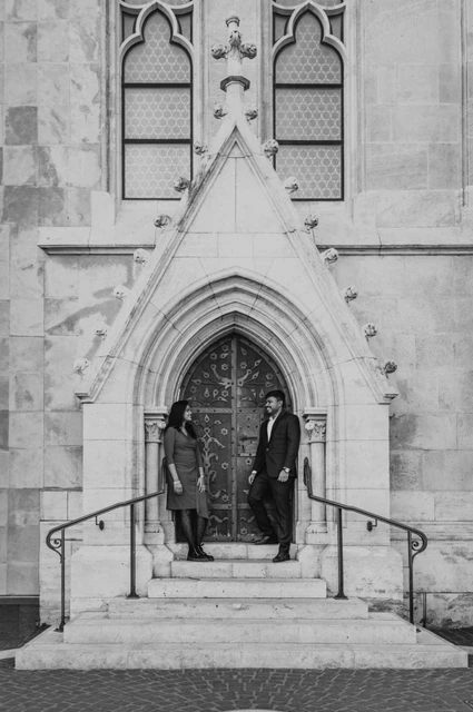 Budapest photoshoot at the Gothic doorway in black and white Black-and-white portrait of the couple standing on the church steps beneath a tall Gothic doorway during their Budapest photoshoot.