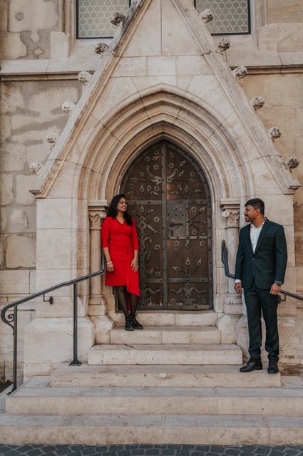 The couple standing apart on stone steps beneath an ornate pointed church doorway during their couple photoshoot in Budapest.