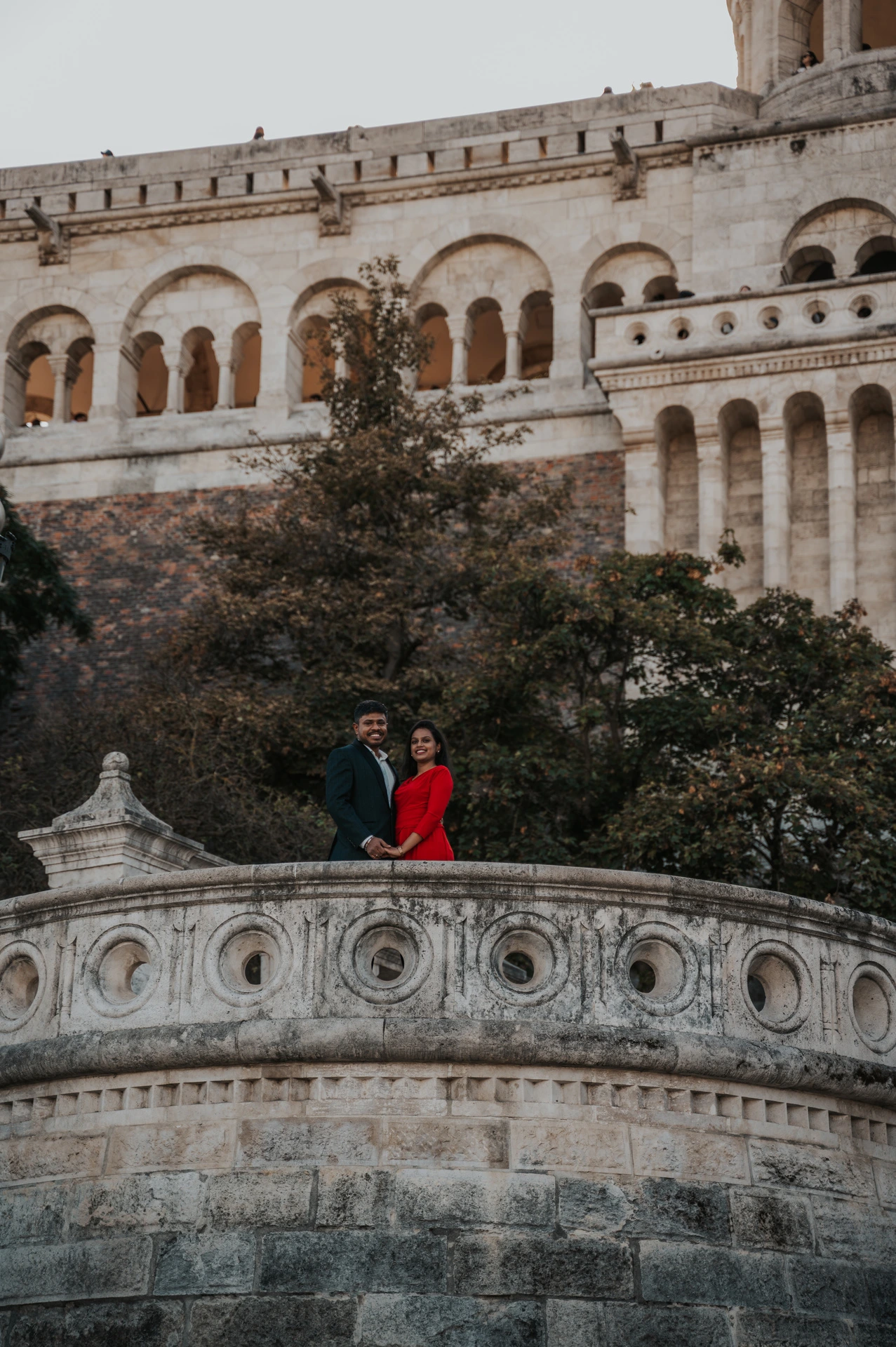 The couple standing behind a carved stone balustrade with trees and Fisherman’s Bastion’s arcades above them during their Budapest couple photoshoot.