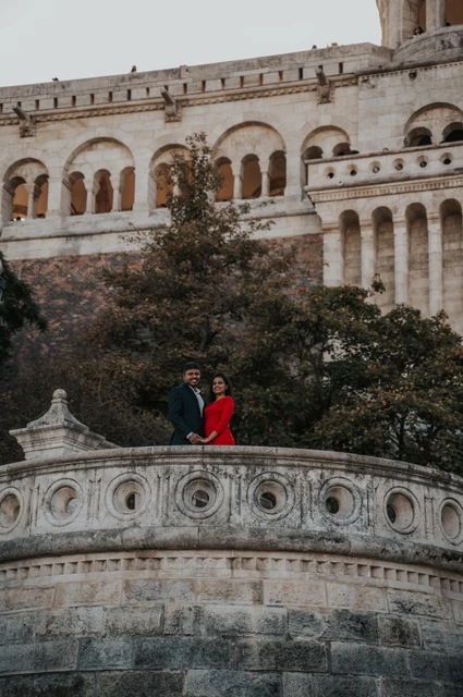 Budapest couple photoshoot by the curved stone balustrade The couple standing behind a carved stone balustrade with trees and Fisherman’s Bastion’s arcades above them during their Budapest couple photoshoot.