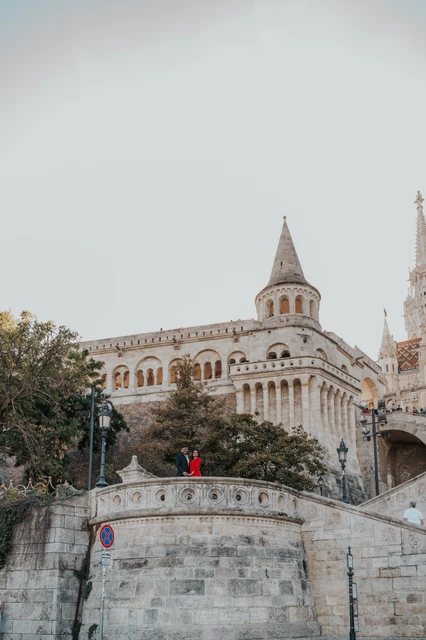 The rounded terrace in a wide Budapest photoshoot view A distant view of the couple on a rounded stone terrace below Fisherman’s Bastion’s tower and arcades during their couple photoshoot in Budapest.