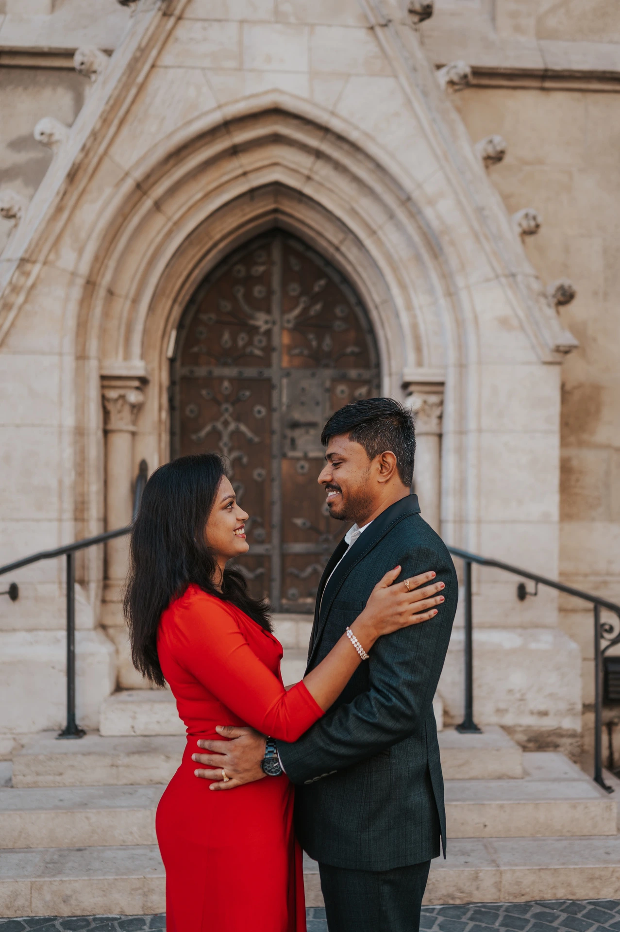 The couple embracing and smiling at one another in front of a carved wooden church door during their couple photoshoot in Budapest.