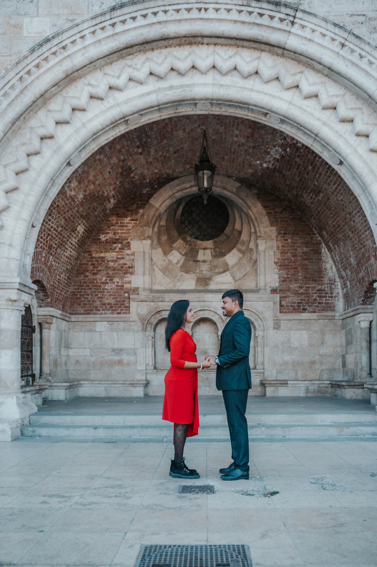The couple facing one another beneath a broad carved arch, framed by stone columns and a brick vault during their Budapest couple photoshoot.