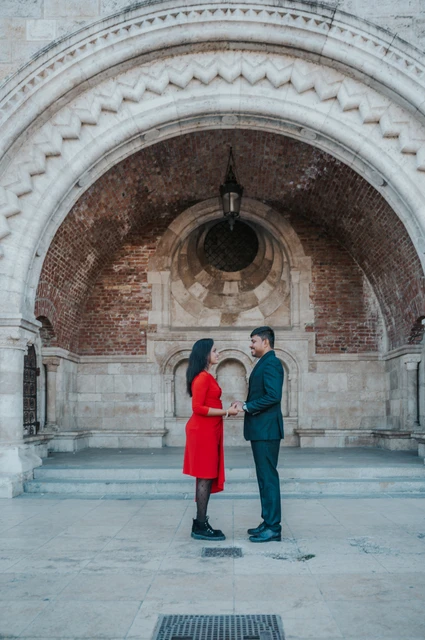 The full stone arch frames a Budapest couple photoshoot The couple facing one another beneath a broad carved arch, framed by stone columns and a brick vault during their Budapest couple photoshoot.