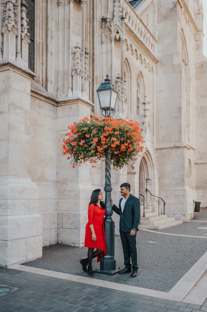 The couple standing beside a lamp-post with hanging red flowers and the pale stone church wall during their photoshoot in Budapest.