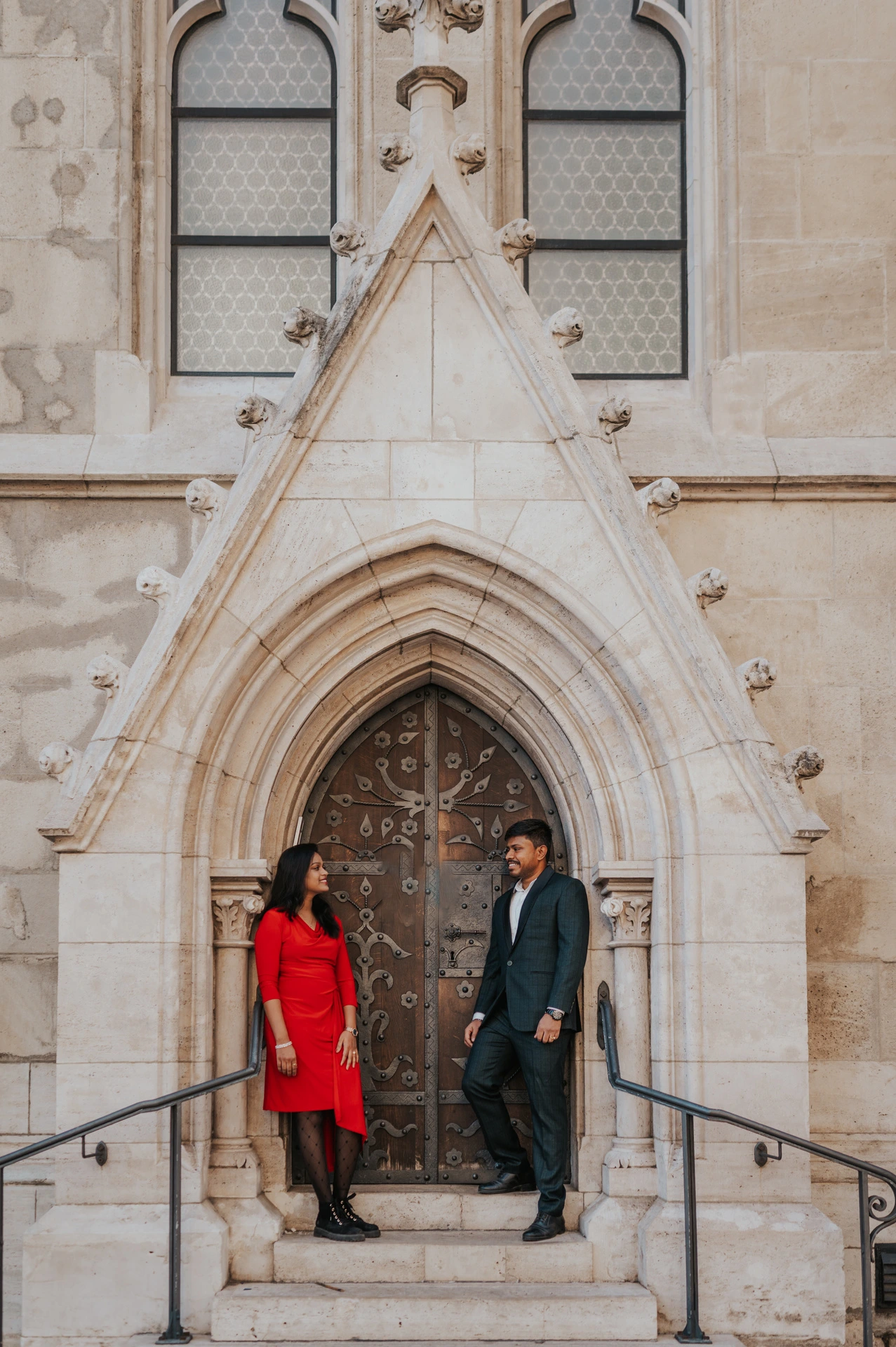 The woman in a red dress and the man in a deep teal suit standing beneath a pointed stone doorway during their Budapest couple photoshoot.