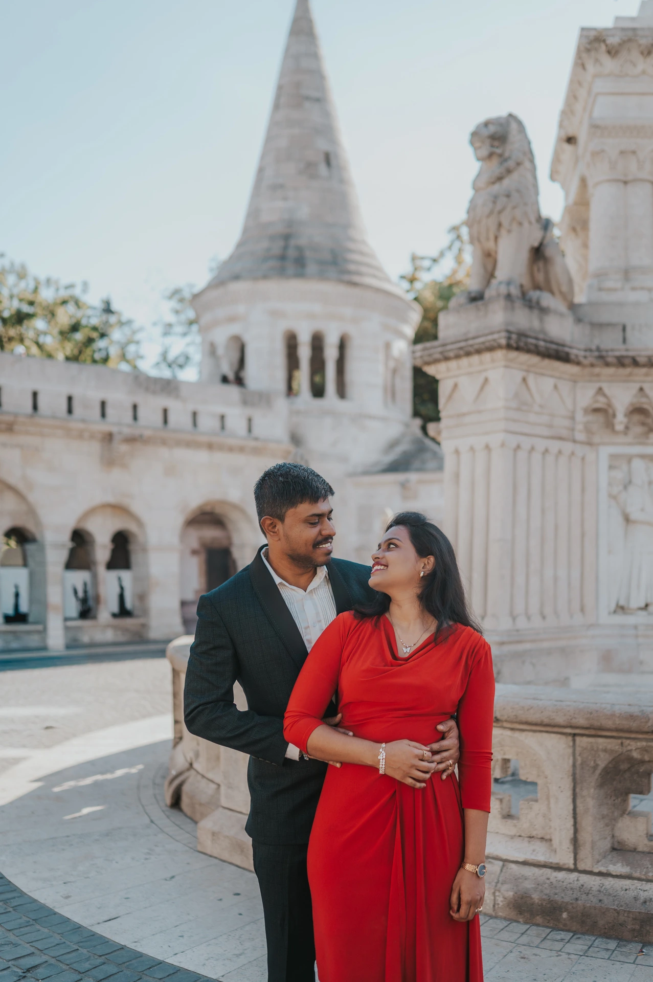 The couple in a red dress and deep teal suit standing together with a pointed turret behind them during their Budapest photoshoot at Fisherman’s Bastion.