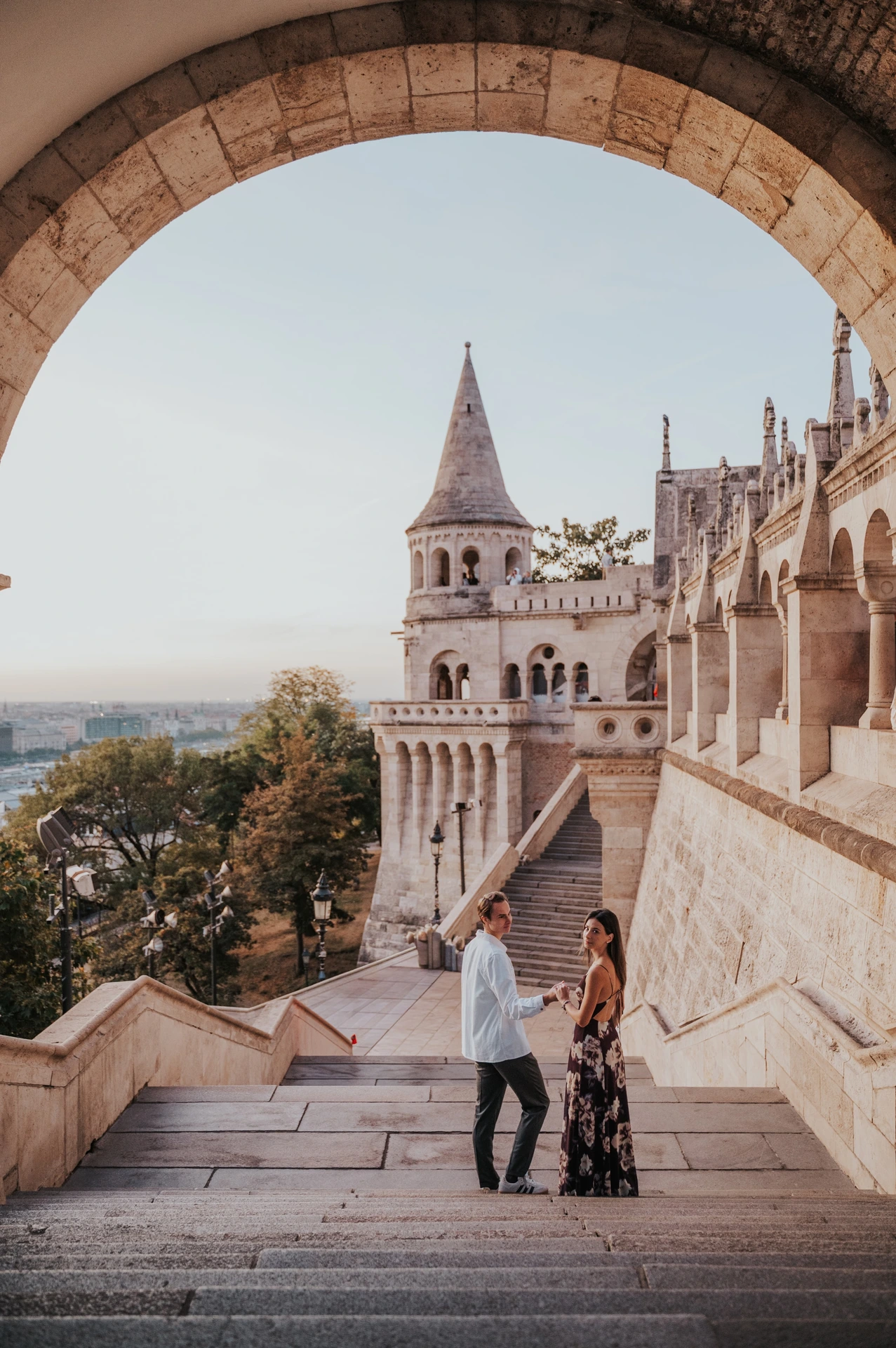 The couple holding hands on the broad stone steps during their Budapest photoshoot, framed by a curved arch.
