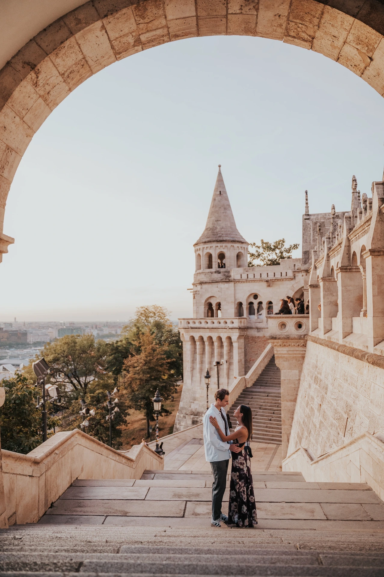 The couple standing close on the staircase with a pointed tower behind them during their Budapest photoshoot.
