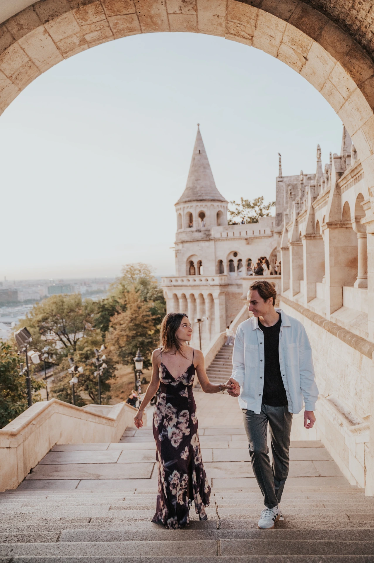 The couple walking up the steps hand in hand during their Budapest photoshoot, her floral dress beside his pale blue shirt.