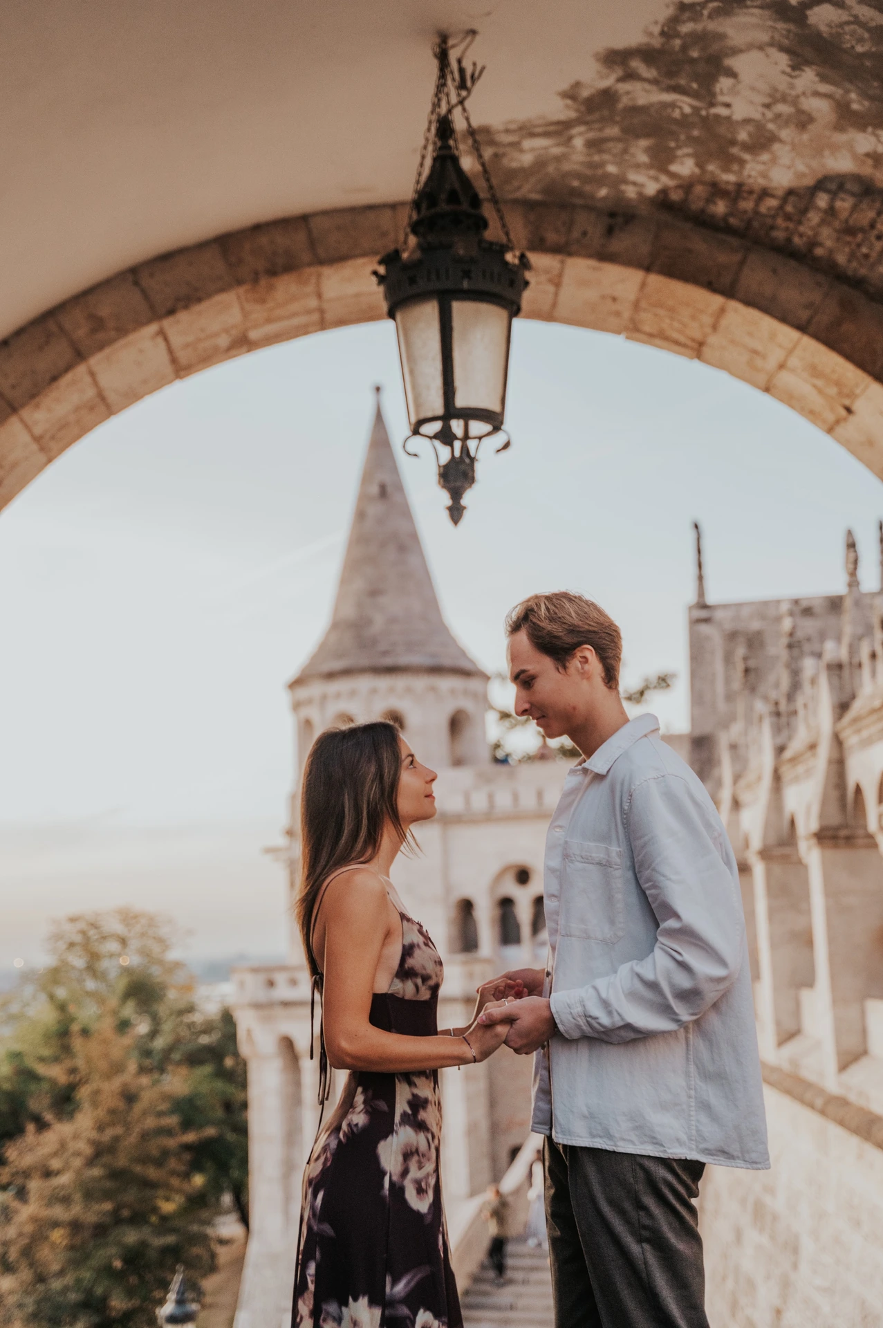 The couple facing each other and holding hands beneath a stone arch during their Budapest photoshoot at Fisherman’s Bastion.