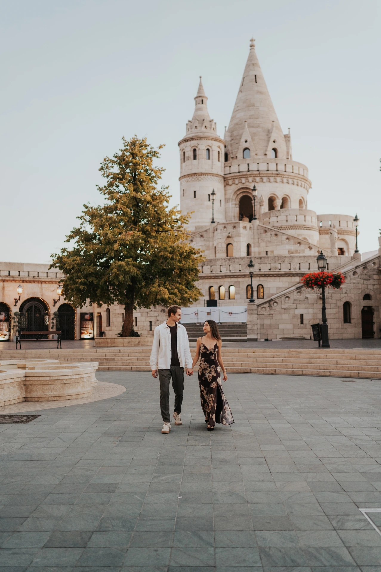 The couple walking together across the square with the pale towers of Fisherman’s Bastion behind them during their Budapest photoshoot.