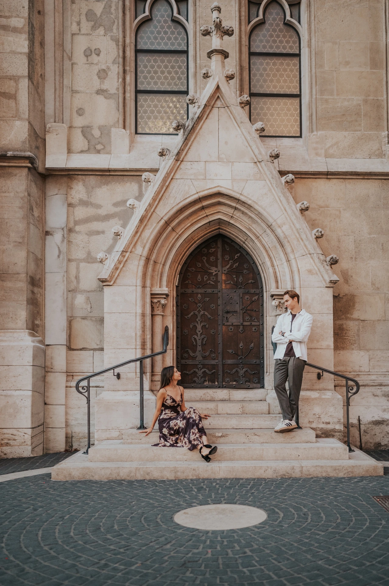 The woman sitting on the steps while her partner stands beside the carved church doorway during their Budapest photoshoot.