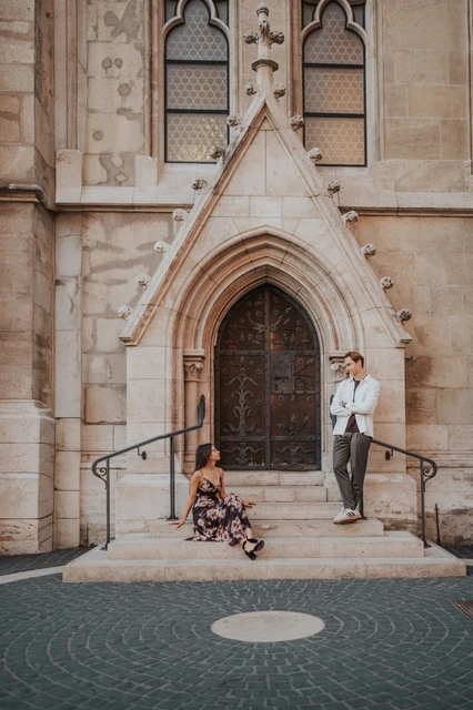Budapest couple photoshoot at a carved church doorway The woman sitting on the steps while her partner stands beside the carved church doorway during their Budapest photoshoot.